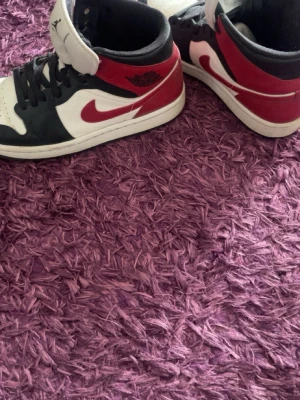 Nike Air Jordan 1 Mid svart/röd/vit - Säljer ett par Nike Air Jordan 1 Mid sneakers i svart, rött och vitt läder. Klassisk design med röd swoosh, svart snörning och perforerad tåbox. Wings-logga på sidan och Jumpman på plösen. Perfekta för dig som gillar streetwear och ikoniska sneakers.