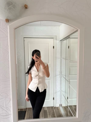 Vit figursydd väst med knappar - Snygg vit blus med v-ringning och knappar framtill. Blusen har en figurnära passform och är ärmlös.