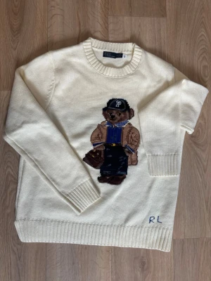 White knitted sweater Polo Bear Ralph Lauren - White knitted sweater from Polo Ralph Lauren with the iconic Polo Bear motif on the front. The sweater has ribbed cuffs and a round neckline. RL embroidery at the bottom. Perfect for those who like classic and playful style.