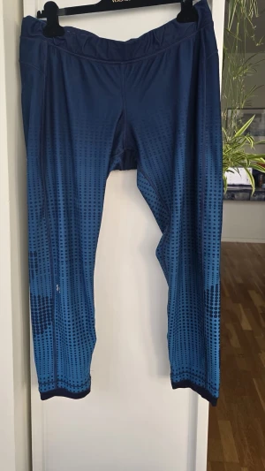 Under armour Blå träningsbyxor size XL - Snygga blå träningsbyxor med svart prickigt mönster. Byxorna har en bred resår i midjan och är tillverkade i ett stretchigt funktionsmaterial som passar perfekt för gym eller löpning. Modellen är 7/8-längd och har en modern, sportig look.något uttöjd i resåren, har ett hål som blivit lagat ( påverkar inte funktionen) 