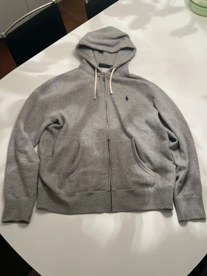 Grå hoodie från Polo Ralph Lauren - Klassisk grå hoodie från Polo Ralph Lauren med dragkedja, två fickor framtill och vit dragsko i huvan. Mörkblå logga broderad på bröstet. Tillverkad i mjukt bomullsmaterial som är riktigt skönt. 