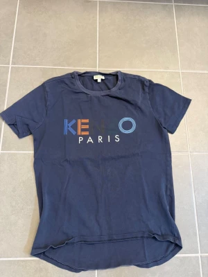 Marinblå Kenzo t-shirt med tryck stl 152 - Snygg marinblå t-shirt från Kenzo med färgglatt logotyptryck på bröstet. Klassisk rund halsringning och korta ärmar. Tillverkad i mjuk bomull som känns skön mot huden. Perfekt för dig som vill ha en stilren men ändå unik look.