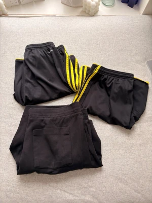 Svarta shorts i bomull  - Tre par svarta shorts, ett par med gula ränder längs sidorna och två utan detaljer. Alla har elastisk midja och fickor, ett par har bakficka. Perfekta för träning eller chill. Materialet känns mjukt och bekvämt.