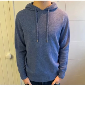 Blå kashmir hoodie - Mysig blå hoodie med huva och dragsko. Tillverkad i mjukt stickat merinoull/kashmir som känns skönt mot huden. Hoodien har långa ärmar och ribbade muddar vid ärmslut och nederkant. Perfekt för en avslappnad och stilren look.