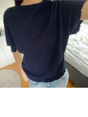 Mörkblå strukturerad topp - En mörkblå topp med korta ärmar och strukturerat tyg. Modellen är loose fit och har en enkel, stilren design utan synliga detaljer. Perfekt att matcha med jeans för en avslappnad look.