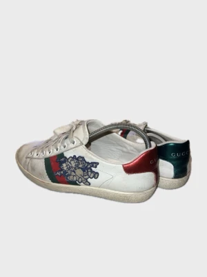 Gucci Ace sneakers  - Vita Gucci Ace sneakers i läder med klassiska gröna och röda ränder på sidan samt broderad blå och beige teddybjörn. Röd metallic-detalj på ena hälen och grön på den andra. Lågt skaft och vita snören. Snygg och ikonisk design.