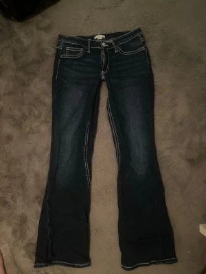 Mörkblå bootcut jeans med kontrastsömmar - ! Eget sytt extremt lågmidjat! Säljer ett par mörkblå bootcut jeans med vita kontrastsömmar och snygga detaljer på bakfickorna. Jeansen har låg midja, klassisk femficksdesign och är tillverkade i stretchigt denimtyg för skön passform. Perfekta för en avslappnad och trendig look.