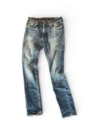 Blå slitna jeans från Nudie Jeans - Säljer ett par blå slitna jeans från Nudie Jeans med klassisk femficksmodell och raka ben. Jeansen har tydliga slitningar och tvättade detaljer för en vintagekänsla. Bakfickorna har Nudie Jeans signaturbrodyr och läderpatch i midjan.  W(30) L (32)