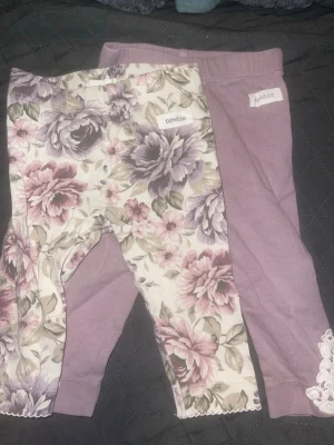  Newbie - Två par leggings från Newbie, ett par i lila och ett par med blommigt mönster i lila, rosa och grönt. Båda har resår i midjan och mjukt bomullsmaterial. Blommiga paret har vågig kant nertill och lila paret har spetsdetalj vid benslut.