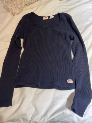 Mörkblå långärmad topp från Levis - Mörkblå ribbad långärmad topp från Levi's med rundad urringning och liten rosett framtill. Diskret Levi's-märke nertill på sidan. Tunn och stretchig bomullskvalitet som passar perfekt till jeans eller kjol.