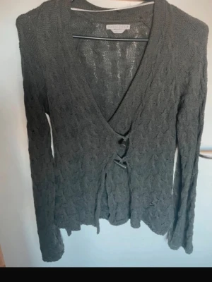 Mörkgrå stickad omlottkofta - Säljer en mörkgrå stickad omlottkofta. Koftan har långa ärmar, knytband framtill och ett diskret kabelstickat mönster. Perfekt för lager på lager och ger en avslappnad men snygg look.