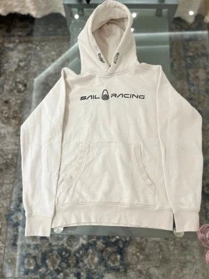 Vit sail racing hoodie - Säljer en stilren vit hoodie från Sail Racing med svart logga på bröstet. Hoodien har klassisk känguruficka, dragsko i huvan och ribbade muddar vid ärmar och nederkant. Perfekt för en avslappnad och sportig look. Som ny 