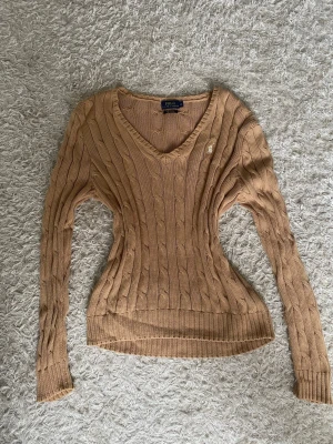 Beige/brun kabelstickad tröja Polo Ralph Lauren - Snygg beige/brun kabelstickad tröja från Polo Ralph Lauren med v-ringning och lång ärm. Tröjan har det klassiska broderade logotypen i vitt på bröstet och ribbade muddar. 