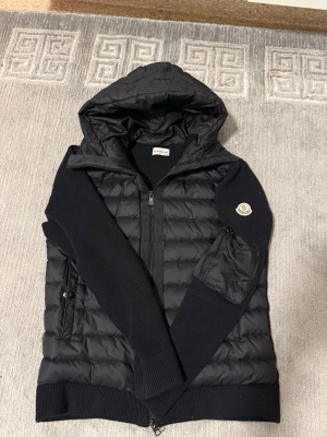 Svart pufferjacka från Moncler - Snygg svart pufferjacka från Moncler med huva och dragkedja framtill. Jackan har stickade ärmar och sidor, quiltad kropp och Moncler-logga på ärmen. Perfekt för kalla dagar och har en modern, sportig look.