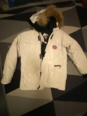 Vit dunjacka från Canada Goose - Säljer en vit dunjacka från Canada Goose med stor huva och pälskant. Jackan har flera praktiska fickor, dragkedja och tryckknappar framtill samt den klassiska Canada Goose-loggan på bröstet. Står xi fast passar L och medium