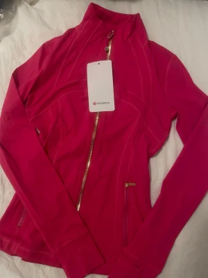 Rosa träningsjacka från Lululemon - Snygg rosa träningsjacka från Lululemon med guldiga dragkedjor och figurnära passform. Jackan har hög krage och långa ärmar, perfekt för att hålla värmen under träning eller när du är på språng. Materialet är mjukt och stretchigt för maximal komfort.