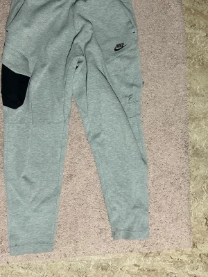 Nike tech byxor - Säljer ett par gråa mjukisbyxor från Nike med svart detaljficka på ena benet och diskret Nike-logga framtill. Byxorna har avsmalnande ben och elastisk midja för skön passform. Perfekta för chill eller träning. Storlek M knapppast använt