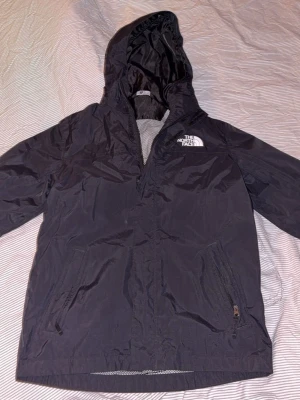 Svart vindjacka från The North Face - Snygg svart vindjacka från The North Face med huva och dragkedja framtill. Jackan har två fickor med dragkedja och meshfoder på insidan. Loggan är tryckt i vitt både fram och bak. Perfekt för blåsiga dagar och enkel att matcha med olika outfits.
