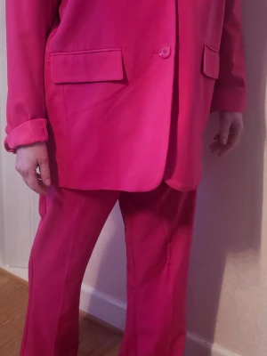 Kostym i stark rosa med kavaj - Säljer en riktigt snygg kostym i stark rosa färg. Kavajen har klassiska fickor med lock och en enkel knappstängning. Byxorna är raka och har hög midja. Perfekt för dig som vill sticka ut och gillar färg! Materialet känns mjukt och bekvämt.