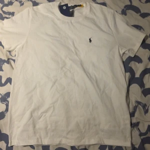 Vit t-shirt från Polo Ralph Lauren - Klassisk vit t-shirt från Polo Ralph Lauren med den ikoniska lilla logotypen broderad på bröstet. T-shirten har rund hals och korta ärmar, tillverkad i mjuk bomull för en skön känsla. Perfekt basplagg till garderoben.
