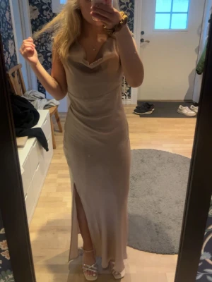 Beige långklänning med slits - Elegant beige långklänning med draperad urringning och hög slits framtill. Klänningen är ärmlös och har en mjuk, följsam passform som ger en stilren siluett. Perfekt för sommarens speciella tillfällen. BARA ANVÄNDA 1 GÅNG! Xs men passar mig som är S/m