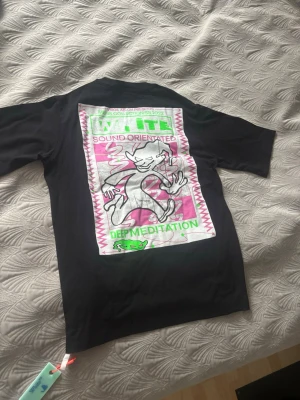 Svart Off-White t-shirt med tryck - Svart t-shirt från Off-White med grönt 'off' tryck framtill och stor grafisk print på ryggen i vitt, rosa och grönt. Klassisk passform och rund hals. Äkta Off-White kommer med original förpackning Storlek S men kan även passa M. Endast testat hemma en gång annars aldrig använt.