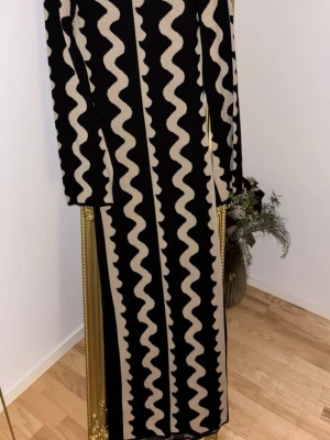 Svart-beige mönstrad kappa NA-KD - Lång kappa från NA-KD i svart med beige vågigt mönster. Rak passform och långa ärmar. Perfekt statement-plagg för dig som gillar unika mönster och vill sticka ut. Tjockare material som passar bra under kallare månader.