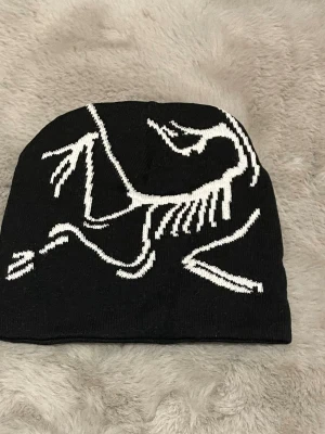 Svart mössa med vit Arc'teryx-logga - Säljer en svart mössa från Arc'teryx med den ikoniska vita dinosaurieloggan i stickat mönster. Mössan är mjuk och stretchig, perfekt för kalla dagar. Materialet är troligtvis akryl eller ullblandning. Snygg och stilren design som sticker ut.