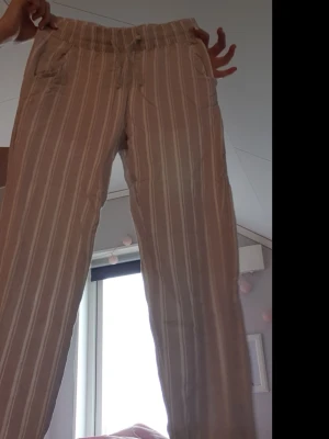 Beige randiga linnebyxor - Snygga beige linnebyxor med vita vertikala ränder och avslappnad passform. Byxorna har resår i midjan, fickor fram och bak samt raka ben. Perfekta för en chill och stilren look. Materialet känns lätt och luftigt, perfekt för varmare dagar.
