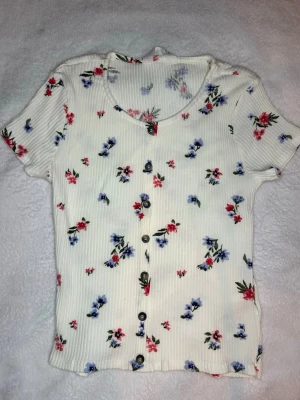 Vit blommig topp med knappar - Vit topp med små blommor i rött och blått. Toppen har korta ärmar, ribbad struktur och knappar framtill. Rund halsringning och mjukt material som känns skönt mot huden.
