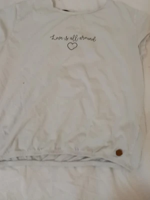 Vit t-shirt med text och hjärta - Säljer en vit t-shirt med texten 'Love is all around' och ett litet hjärta tryckt framtill. T-shirten har korta ärmar och en avslappnad passform. Perfekt till vardags för en enkel och söt stil.