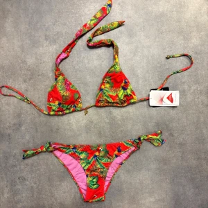 Färgglad bikini med tropiskt mönster - Snygg trekantsbikini från PHAX med tropiskt mönster. Både överdel och underdel har knytband i sidorna och nacken. Överdel är lite vadderad. Detaljer både på över-och nederdel. Har dock klippt bort lappen i badtrosorna (är ofta för snabb att klippa bort lappar, utan att det används) Men både över-och underdel är såklart helt oanvända! Alla lappar är kvar på överdelen. Är en storlek S, men eftersom man kan justera båda delar, så passar det även mindre och lite större storlekar.