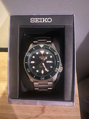 Seiko automatisk klocka i stål - Snygg automatisk klocka från Seiko med grön urtavla och vridring, samt dag- och datumvisning. Klockan har ett robust armband i silverfärgat stål och levereras i originalask med extra länkar och manual. Köpt för 4999