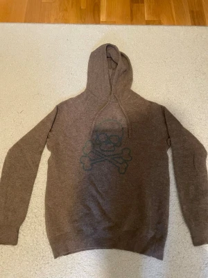 Brun hoodie med dödskalletryck - Säljer en brun hoodie från Scalpers  med ett stort dödskalle- och korsade benknotor-tryck på bröstet. Hoodien har dragsko i huvan och är tillverkad av kashmir. 