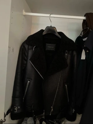 Svart skinnjacka från Oakwood - Snygg svart skinnjacka från Oakwood med bred krage i fuskpäls, dragkedjor på fickor och ärmslut samt bältesdetaljer vid midjan. Jackan har en cool bikerstil och är fodrad för extra värme. Perfekt för dig som vill ha en stilren och trendig look.