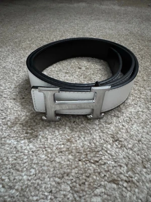 Vitt skärp med H-spänne - Snyggt vitt skärp från H-bälte med ett stort, silverfärgat H-spänne. Bältet har en stilren och klassisk design med svart insida och är tillverkat i skinn. Perfekt accessoar för att lyfta vilken outfit som helst. Helt oanvänt.