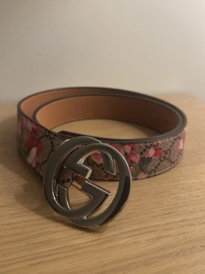Gucci blommigt bälte med GG-spänne - Snyggt bälte från Gucci med klassiskt GG-spänne i silverfärg. Bältet har ett blommigt mönster i rosa och rött på brun bakgrund med GG-loggor. Tillverkat i skinn med exklusiv känsla och passar perfekt för att lyfta vilken outfit som helst.