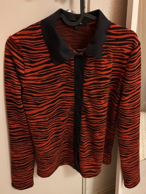 Röd och svart tiger-mönstrad kofta - Snygg långärmad kofta med röd och svart tiger-mönster, mörkgrå krage och knappslå framtill. Materialet känns mjukt och stretchigt. Oanvänd.