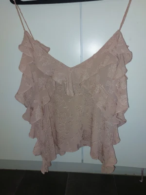 Beige volangtop med tunna axelband - Supersöt rosa linnetopp med volanger och tunna axelband. Toppen har ett blommigt mönster i tyget och en luftig, flowy passform. Perfekt för dig som gillar romantisk och feminin stil.