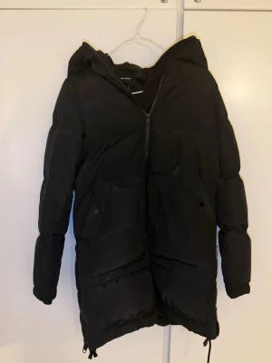 Svart pufferjacka med huva - Säljer en svart pufferjacka med huva och dragkedja framtill. Jackan har två stora fickor med knappstängning och justerbar dragsko nedtill. Perfekt för kalla dagar och riktigt skön att bära. Enkel, stilren och passar till det mesta.