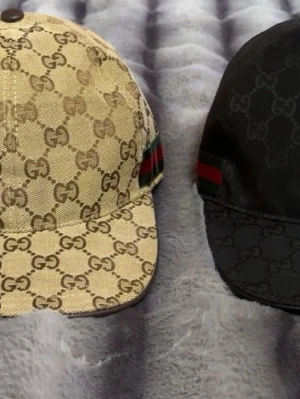 Gucci keps med GG-mönster - Säljer en snygg Gucci keps med det klassiska GG-monogrammet över hela kepsen. Finns i beige, svart och grått tyg med den ikoniska gröna och röda randen på sidan. Justerbar baktill och tillverkad i canvasmaterial. Perfekt för dig som vill ha en exklusiv och trendig look.