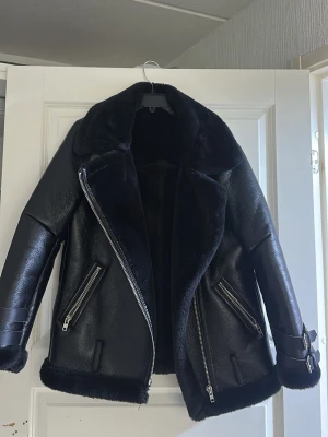 Svart pälsjacka med dragkedjor - Snygg svart pälsjacka i bikerstil med stora slag, fuskpälsfoder och detaljerade silverfärgade dragkedjor. Jackan har bälte vid ärmsluten och två snedställda fickor framtill. Perfekt för kalla vinterdagar och ger en cool look.