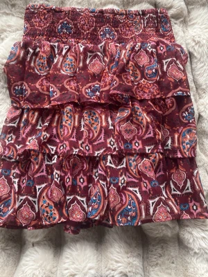 Mönstrad kortkjol med volanger - Säljer en kort kjol med paisleymönster i vinröda och rosa toner, inslag av blått och vitt. Kjolen har flera lager med volanger och ett smockat midjeband som ger en skön passform. Materialet känns lätt och luftigt, perfekt för varmare dagar.