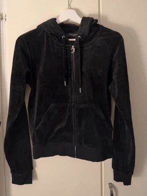 Svart velourhoodie från Juicy Couture - Mjuk och lyxig svart velourhoodie från Juicy Couture med dragkedja framtill, huva med snören och broderad logga på bröstet. Klassisk känguruficka och ribbade muddar vid ärmslut och nederkant. Perfekt för en chill och trendig look.