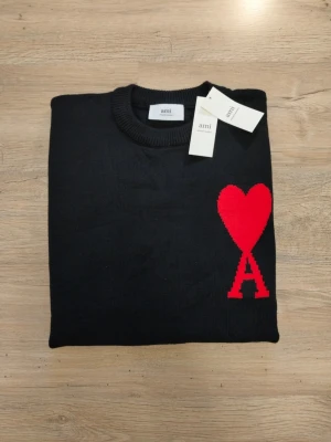 Svart stickad tröja från Ami Paris - Svart stickad tröja från Ami Paris med rund halsringning och ribbade muddar. På bröstet finns ett stort rött hjärta med bokstaven A. Tröjan har en klassisk passform och är tillverkad i mjukt material. Perfekt för dig som gillar stilrena detaljer.