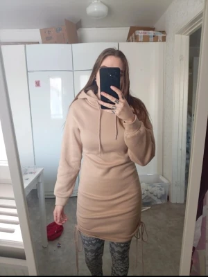 Beige hoodieklänning med snörning - Cool beige hoodieklänning med snörning i sidorna och huva. Klänningen är kort och har långärmar samt en relaxed passform. Perfekt för dig som gillar streetstyle och vill ha något bekvämt men ändå snyggt.