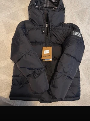 Svart pufferjacka från Burberry - Snygg svart pufferjacka från Burberry med huva och dragkedja framtill. Jackan har logotypen 'BURBERRY LONDON ENGLAND' på ärmen och två praktiska fickor med dragkedja. Perfekt för kalla dagar och har en modern, boxig passform.