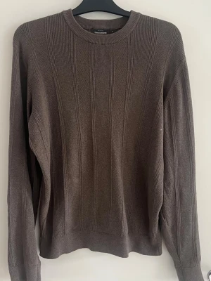 Brun stickad tröja från Dressmann - Säljer en brun stickad tröja från Dressmann i storlek M. Tröjan har rund halsringning, ribbade muddar och ett diskret vertikalt mönster. Perfekt för dig som gillar en enkel och stilren look.