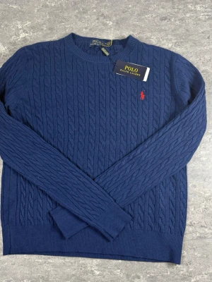 Marinblå kabelstickad tröja Polo Ralph Lauren - Kabelstickad marinblå tröja från Polo Ralph Lauren med rund halsringning och röd broderad logga på bröstet. Tröjan har långa ärmar och ribbade muddar vid ärmslut och nederkant. Klassisk och stilren design som passar till mycket.