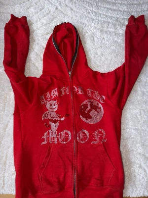 Röd hoodie med tryck och dragkedja - Säljer en röd hoodie med dragkedja och huva. På framsidan finns ett tryck med texten 'AIM FOR THE MOON',🌕 en tecknad figur och en måne i glittriga detaljer. Hoodien har långa ärmar och fickor framtill. Perfekt för dig som gillar streetwear och vill sticka ut. Man får även med en gratis vara på bilden i slutet🔥☺️